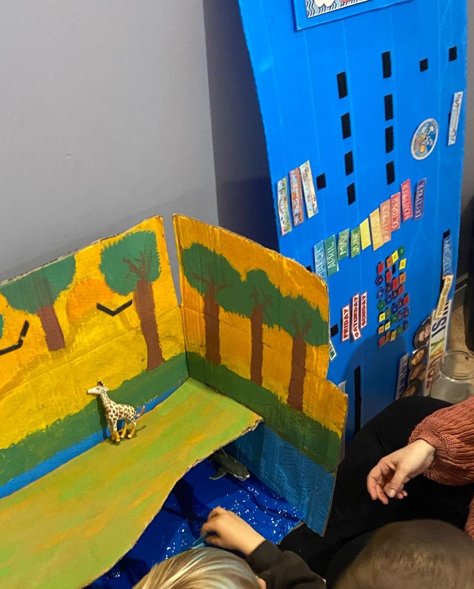 A hand-painted cardboard savanna habitat with a toy giraffe, used to learn about animal habitats in English, next to a blue English class calendar
